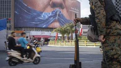 An armed Iranian police officer holding a rifle monitors the area as motorcyclists ride beneath a billboard depicting an AI-generated image of the Strait of Hormuz and an effigy of US President Donald Trump, displayed on the wall of a state building in downtown Tehran, Iran, on May 3, 2026.