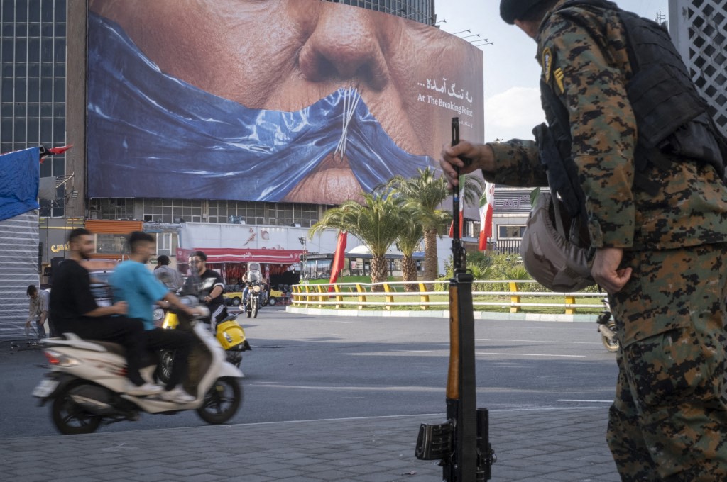 An armed Iranian police officer holding a rifle monitors the area as motorcyclists ride beneath a billboard depicting an AI-generated image of the Strait of Hormuz and an effigy of US President Donald Trump, displayed on the wall of a state building in downtown Tehran, Iran, on May 3, 2026.
