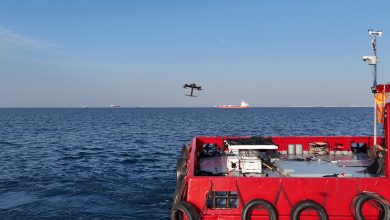 A quadcopter drone hovers over open water ahead of a red vessel's deck, illustrating the UK's development of an automatic drone landing platform for moving ships